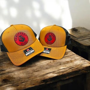 May include: Two brown and black trucker hats with a red and black logo that says "Homefront Stix" on the front. The hats are on a wooden surface.