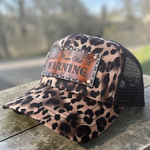 May include: Leopard print trucker hat with a brown leather patch that reads "Should've Come With A WARNING". The hat has a black mesh back and a curved brim. The hat is sitting on a wooden surface.