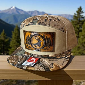 May include: A camouflage print flat bill snapback hat with a tan mesh back and a tan front panel. The front panel features a leather patch with a deer silhouette inside a circle with trees. The hat has a Richardson tag.