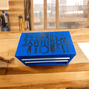 May include: A blue metal toolbox with two drawers, displaying the name "JARRISON" and tool illustrations. The toolbox is placed on a wooden surface. The background shows a wooden wall and a window.
