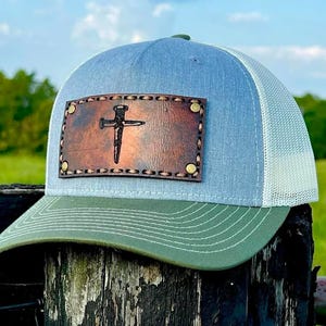 May include: A blue and white trucker hat with a green brim. The hat has a brown leather patch with a nail hammered into a cross design.