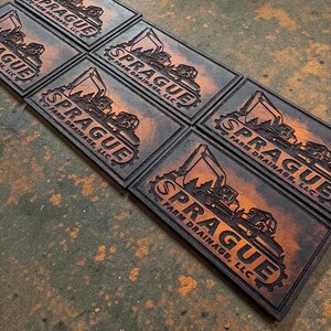 May include: Five rectangular patches featuring a dark brown and orange gradient finish. Each patch displays an embossed graphic of an excavator and the text "SPRAGUE FARM DRAINAGE, LLC" arranged in a gear-shaped border design.