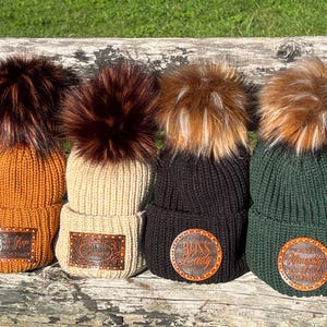 May include: Four knitted beanies with faux fur pom-poms. The beanies are in shades of orange, beige, black, and green. Each beanie has a leather patch with text. The beanies are displayed on a wooden surface.