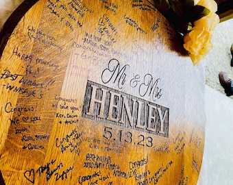 Pode incluir: Um barril de madeira com uma inscrição personalizada "Mr. & Mrs. Henley 5.13.23" e coberto de votos escritos à mão para o casal recém-casado.
