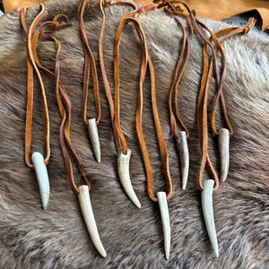 May include: A collection of handmade necklaces featuring off-white antler pendants suspended from brown leather cords. The pendants vary in size and shape, and the cords are knotted at the top. The necklaces are displayed on a textured, brown fur-like surface.