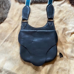 May include: A dark brown leather pouch featuring a curved flap closure and white contrast stitching. The pouch is attached to two adjustable straps that are teal blue with white edges, connected by metal rings to the leather body.