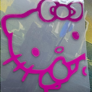May include: A magenta vinyl decal featuring the outline of a cartoon cat character with a bow on its head. The design is presented on a clear adhesive backing, showcasing the bold, vibrant pink lines that define the iconic facial features.
