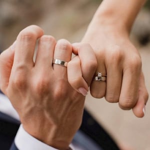 May include: Two hands holding each other, with a simple silver band on the ring finger of the left hand and a diamond ring on the ring finger of the right hand.