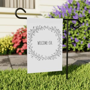 Puede incluir: Una bandera de jardín blanca con un diseño de corona negra y el texto "WELCOME-ISH". La bandera está unida a un soporte de bandera de jardín de metal negro.