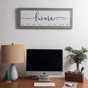 May include: A wooden sign with the word "home" in cursive, EST. 2020, and coordinates. The sign hangs above a desk with a computer, lamp, and decorative items. The desk is made of wood.