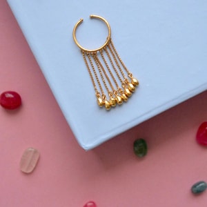 May include: Gold ear cuff with a dangling chain of small, round beads. The cuff is on a light blue surface.
