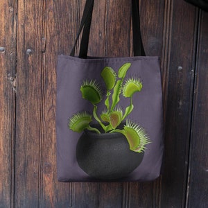 Puede incluir: Un bolso tote de color gris oscuro con una ilustración gráfica de una planta carnívora Venus atrapamoscas verde que crece en una maceta negra redondeada. El bolso tiene dos asas de tela negras y una forma rectangular.