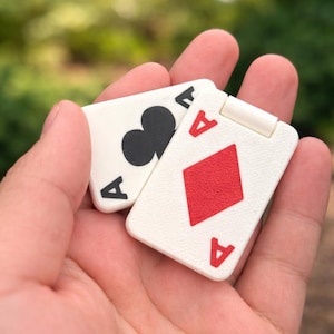 Può includere: Due carte da gioco bianche, l'Asso di Picche e l'Asso di Quadri, sono tenute in una mano. L'Asso di Picche ha un simbolo di picche nero e l'Asso di Quadri ha un simbolo di quadri rosso. Entrambe le carte hanno una "A" rossa negli angoli.