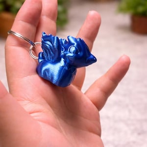 May include: A blue dragon keyring with a silver ring. The dragon figurine has wings and a detailed design. The keyring is made of a shiny, metallic-looking material. The dragon is a vibrant shade of blue.