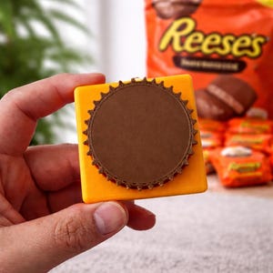 Puede incluir: Un objeto cuadrado y amarillo con un diseño circular marrón en el centro. El objeto tiene un borde decorativo marrón. Una bolsa de Reese's está en el fondo.