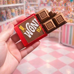 May include: A chocolate bar with a bite taken out, featuring the text "Wonka" on the red wrapper. The chocolate is a rich brown color, and the wrapper has gold accents. The background is a candy store with shelves of colorful treats.