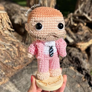 Puede incluir: Una muñeca amigurumi de crochet que lleva un traje rosa con una camisa blanca y una corbata a rayas blancas y negras. La muñeca está de pie sobre una base de madera con el texto "Sheepy Creations Co." grabado en ella.