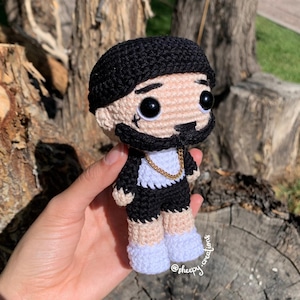 May include: Crocheted amigurumi doll with black hair, a black and white outfit, and a gold chain. The doll is wearing white shoes and has a black beard. The doll is being held in a hand.