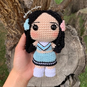 Könnte beinhalten: Gehäkelte Puppe mit schwarzen Haaren, blau-weißem Cheerleader-Outfit und rosa Blumen auf dem Kopf. Die Puppe hält einen Pompon in ihrer rechten Hand. @sheepy_creations ist im Hintergrund geschrieben.