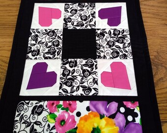 May include: A quilted table runner with a black border. The runner features floral patterns with pink, yellow, and purple flowers, and black and white heart and swirl designs. The center has a black square.