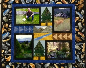 May include: A handmade quilt with a black background adorned with feather patterns. The quilt features a central panel with framed landscape photos and geometric designs in blue, green, and brown. The border is a dark blue.