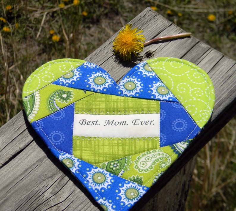May include: A heart-shaped patchwork quilt in shades of blue and green with a white label that reads "Best. Mom. Ever."