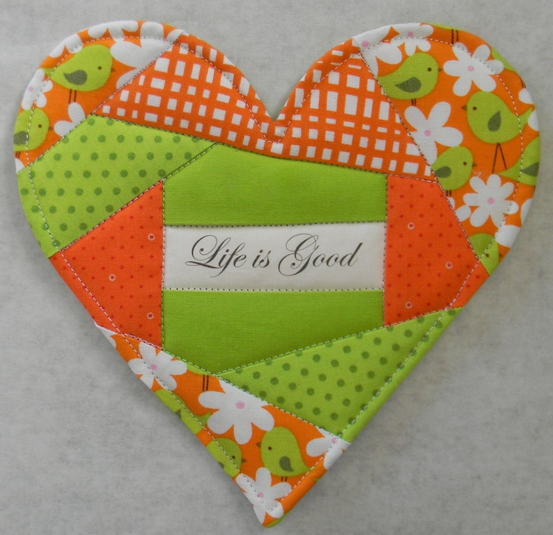 May include: A green and orange patchwork heart-shaped hot pad with the text "Life is Good!" in a white rectangle.