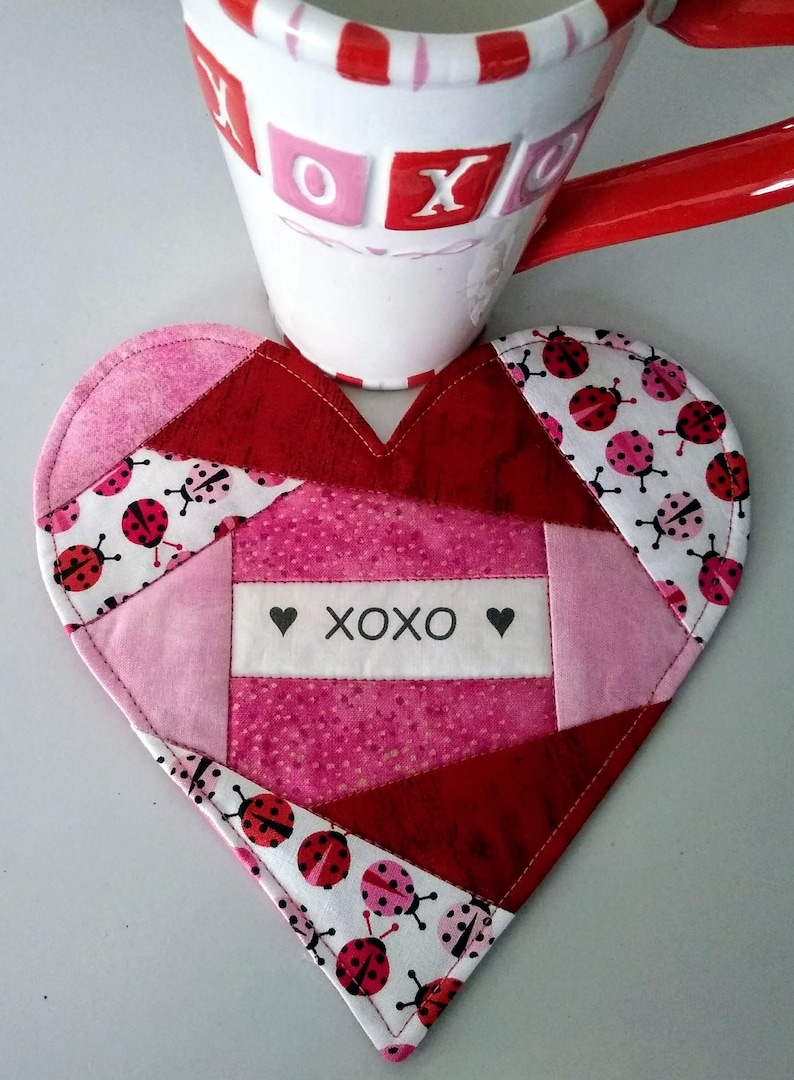 May include: A heart-shaped quilted coaster with a patchwork design in shades of pink and red. The coaster features a white fabric panel with the text "XOXO" and two small hearts. The fabric is decorated with a pattern of ladybugs.
