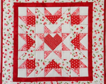May include: A red and white patchwork quilt with a cherry print border. The quilt is made up of squares and triangles, and features a large heart in the center.