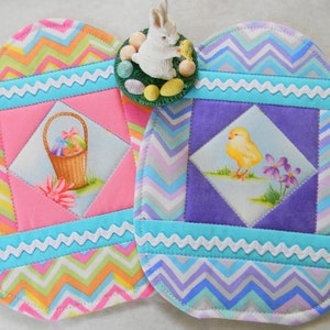 May include: Two handmade quilted Easter egg-shaped hot pads. One features a pink and white chevron pattern with a basket of Easter eggs. The other features a purple and blue chevron pattern with a yellow chick. Both hot pads have a white scalloped trim.
