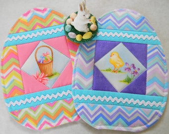 May include: Two handmade quilted Easter egg-shaped hot pads. One features a pink and white chevron pattern with a basket of Easter eggs. The other features a purple and blue chevron pattern with a yellow chick. Both hot pads have a white scalloped trim.
