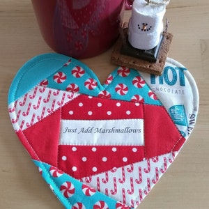 May include: A red and turquoise heart-shaped mug rug with a white fabric panel that says "Just Add Marshmallows". The mug rug is made from patchwork fabric with candy cane and peppermint designs.