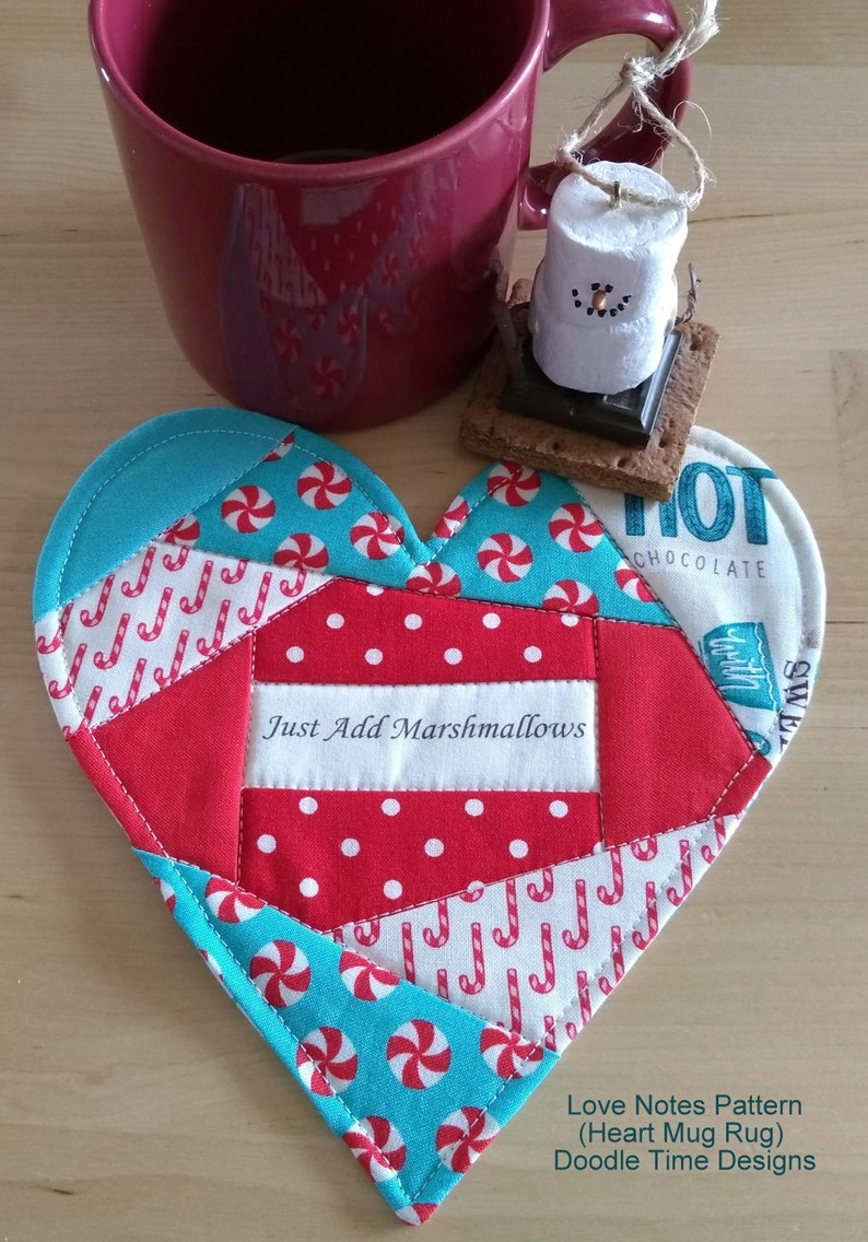 May include: A red and turquoise heart-shaped mug rug with a white fabric panel that says "Just Add Marshmallows". The mug rug is made from patchwork fabric with candy cane and peppermint designs.