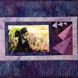 Pode incluir: Uma tapeçaria de parede acolchoada de cor roxa e branca com uma imagem emoldurada de uma estátua de jardim de um anjo rodeado de flores cor-de-rosa. A moldura é rodeada por um design de patchwork roxo e branco.