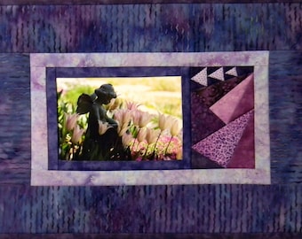 May include: A purple and white quilted wall hanging with a framed picture of a garden statue of an angel surrounded by pink flowers. The frame is surrounded by a purple and white patchwork design.