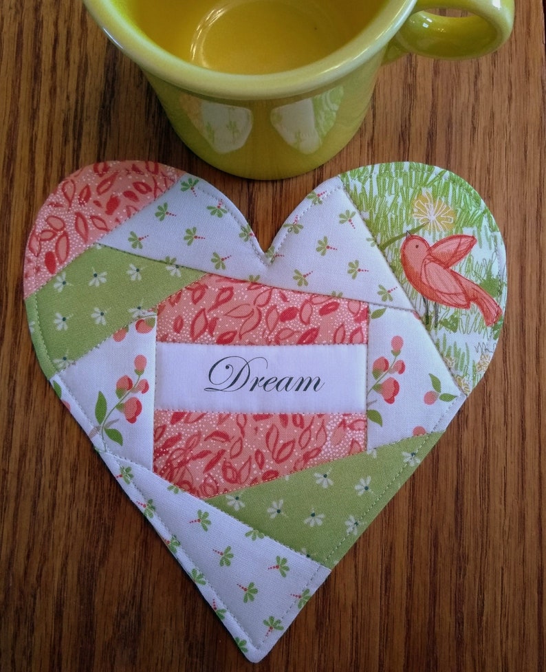 May include: A heart-shaped quilted coaster with a patchwork design in shades of green, white, and pink. The coaster features a small bird applique and the word "Dream" in black lettering.