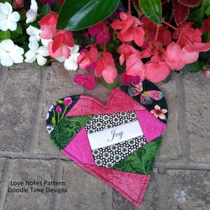 May include: A pink and green patchwork heart-shaped fabric with the word "Joy" written on a white label. The heart is surrounded by pink flowers.