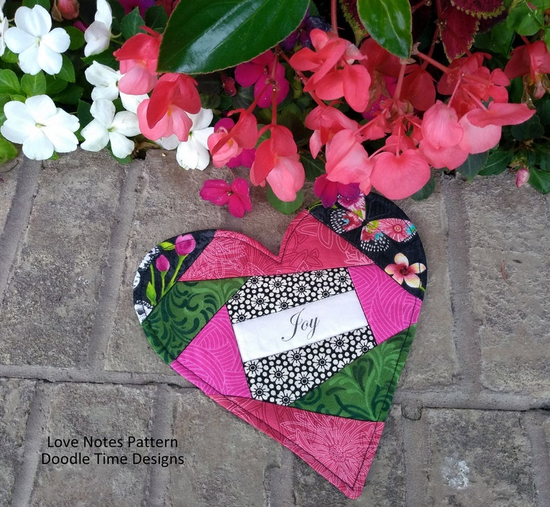 May include: A pink and green patchwork heart-shaped fabric with the word "Joy" written on a white label. The heart is surrounded by pink flowers.