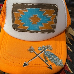 May include: A white trucker hat with an orange mesh back. The front of the hat features a hand-painted design of a brown, orange, and blue geometric pattern. The brim of the hat is orange and has a hand-painted design of two crossed arrows with a blue feather.