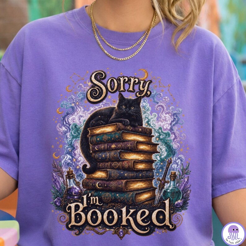 K&ouml;nnte beinhalten: Ein lavendelfarbenes T-Shirt mit einer Grafik einer schwarzen Katze, die auf einem Stapel alter B&uuml;cher liegt. Der Text lautet Sorry, I'm BOOKED. Das Design zeigt wirbelnden violetten und blauen Rauch, Kristalle, einen Dolch und Fl&auml;schchen.