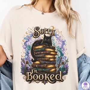 K&ouml;nnte beinhalten: Ein cremefarbenes T-Shirt mit einer Grafik einer schwarzen Katze, die auf einem Stapel alter B&uuml;cher sitzt. Das Design zeigt wirbelnde violette und blaue kosmische Wolken, Zaubertrankflaschen, ein Schwert und den Text Sorry I'm BOOKED in Gold.