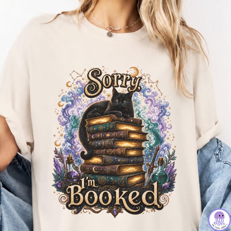 K&ouml;nnte beinhalten: Ein cremefarbenes T-Shirt mit einer Grafik einer schwarzen Katze, die auf einem Stapel alter B&uuml;cher sitzt. Das Design zeigt wirbelnde violette und blaue kosmische Wolken, Zaubertrankflaschen, ein Schwert und den Text Sorry I'm BOOKED in Gold.