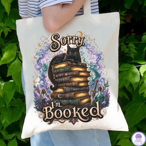 K&ouml;nnte beinhalten: Eine cremefarbene Stofftasche mit einem Grafikdruck einer schwarzen Katze, die auf einem Stapel alter B&uuml;cher sitzt. Der Text lautet Sorry, I'm BOOKED. Das Design zeigt wirbelnde violette und blaue Wolken, Sterne, Kristalle und ein Schwert.