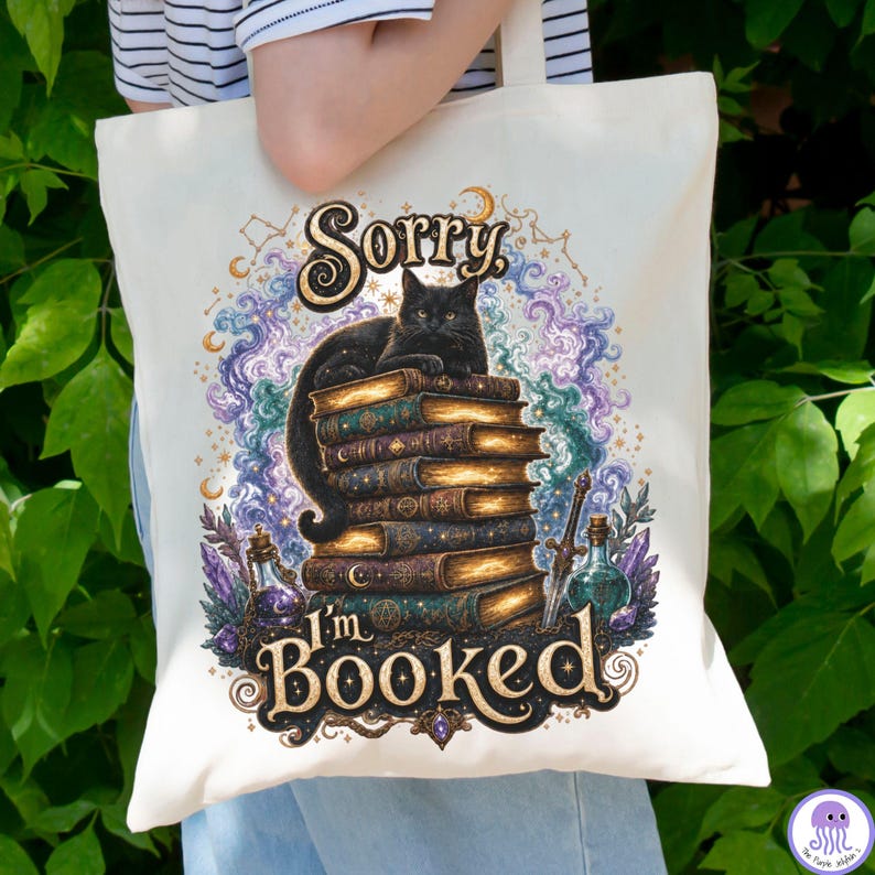 K&ouml;nnte beinhalten: Eine cremefarbene Stofftasche mit einem Grafikdruck einer schwarzen Katze, die auf einem Stapel alter B&uuml;cher sitzt. Der Text lautet Sorry, I'm BOOKED. Das Design zeigt wirbelnde violette und blaue Wolken, Sterne, Kristalle und ein Schwert.