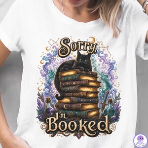 K&ouml;nnte beinhalten: Ein wei&szlig;es T-Shirt mit einem Grafikdruck, der eine schwarze Katze auf einem Stapel alter B&uuml;cher zeigt. Das Design umfasst wirbelnde violette und blaue Wolken, Kristalle, einen kleinen Dolch und den Text Sorry I'm BOOKED in goldener Schrift.