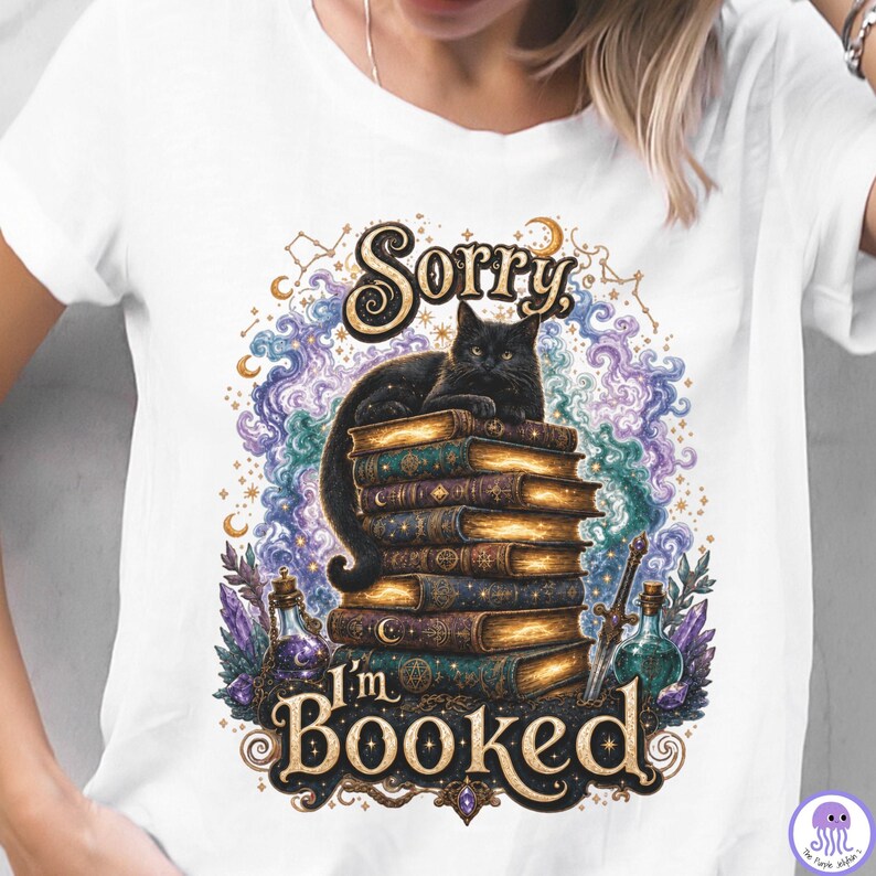 K&ouml;nnte beinhalten: Ein wei&szlig;es T-Shirt mit einem Grafikdruck, der eine schwarze Katze auf einem Stapel alter B&uuml;cher zeigt. Das Design umfasst wirbelnde violette und blaue Wolken, Kristalle, einen kleinen Dolch und den Text Sorry I'm BOOKED in goldener Schrift.