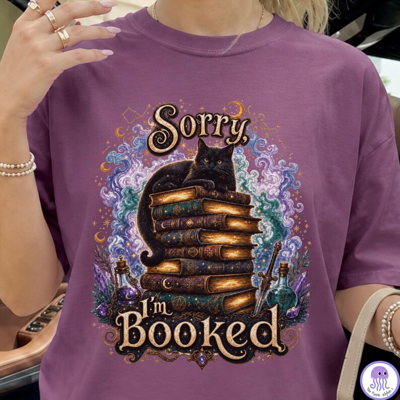 K&ouml;nnte beinhalten: Ein violettes T-Shirt mit einer Grafik einer schwarzen Katze, die auf einem Stapel alter B&uuml;cher sitzt. Der Text lautet Sorry I'm BOOKED. Das Design enth&auml;lt wirbelnden violetten und blauen Rauch, Sterne, Mondsicheln, ein Schwert und zwei Glasflaschen.