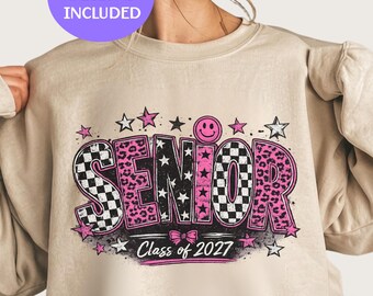 Könnte beinhalten: Ein beigefarbenes Sweatshirt mit Rundhalsausschnitt und einem Grafikdruck des Wortes SENIOR in großen Buchstaben. Die Buchstaben haben schwarz-weiße Karomuster, rosa Leopardenmuster und schwarze Sterne. Enthält ein rosa Smiley, Sterne, eine Schleife und Class of 2027.