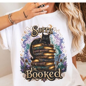 K&ouml;nnte beinhalten: Ein wei&szlig;es T-Shirt mit einer Grafik, die eine schwarze Katze auf einem Stapel alter B&uuml;cher zeigt. Das Design enth&auml;lt wirbelnden violetten und blauen Rauch, Kristalle, Zaubertrankflaschen und den Text "Sorry I'm BOOKED" in goldener Schrift.