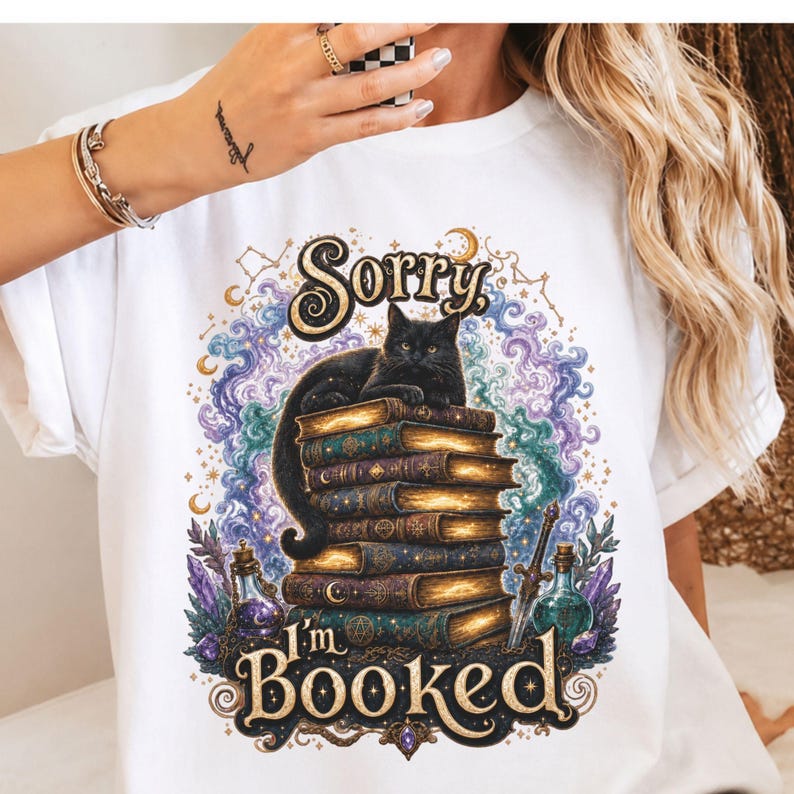 K&ouml;nnte beinhalten: Ein wei&szlig;es T-Shirt mit einer Grafik, die eine schwarze Katze auf einem Stapel alter B&uuml;cher zeigt. Das Design enth&auml;lt wirbelnden violetten und blauen Rauch, Kristalle, Zaubertrankflaschen und den Text "Sorry I'm BOOKED" in goldener Schrift.